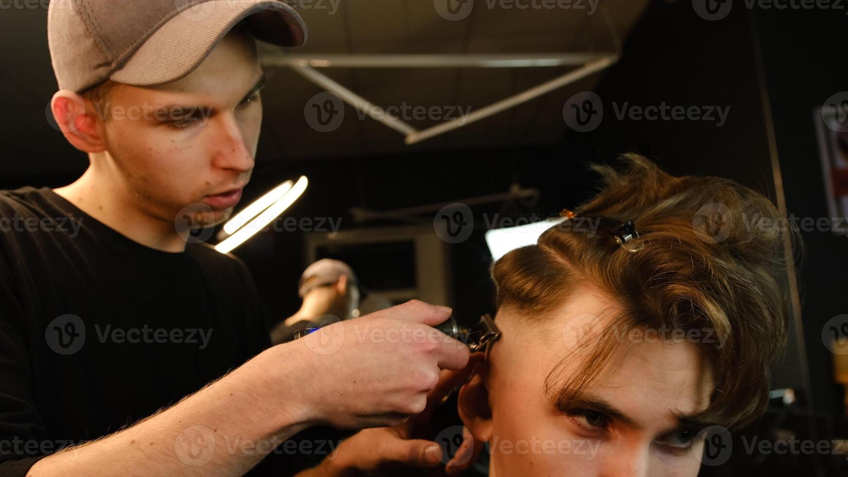 Shot of a handsome barber giving a haircut to his client using trimmer. Hairdresser service in a modern barbershop in a dark key lightning with warm light side view photo