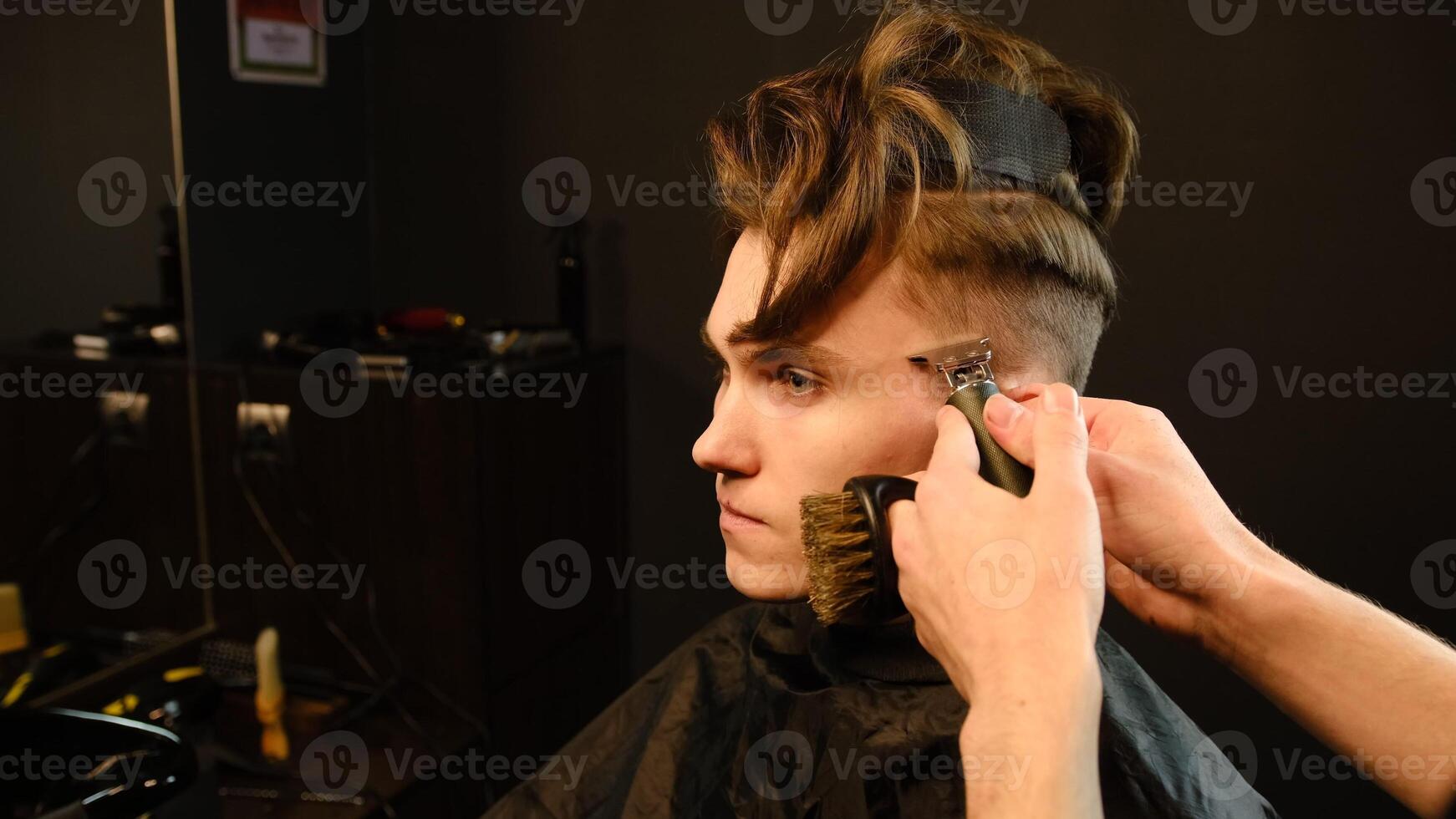 Shot of a handsome barber giving a haircut to his client using trimmer. Hairdresser service in a modern barbershop in a dark key lightning with warm light side view photo