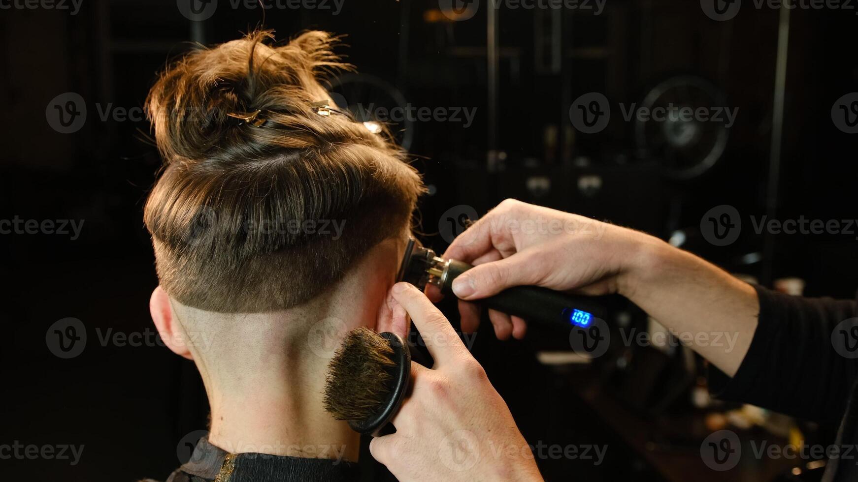 Shot of a handsome barber giving a haircut to his client using trimmer. Hairdresser service in a modern barbershop in a dark key lightning with warm light back view photo