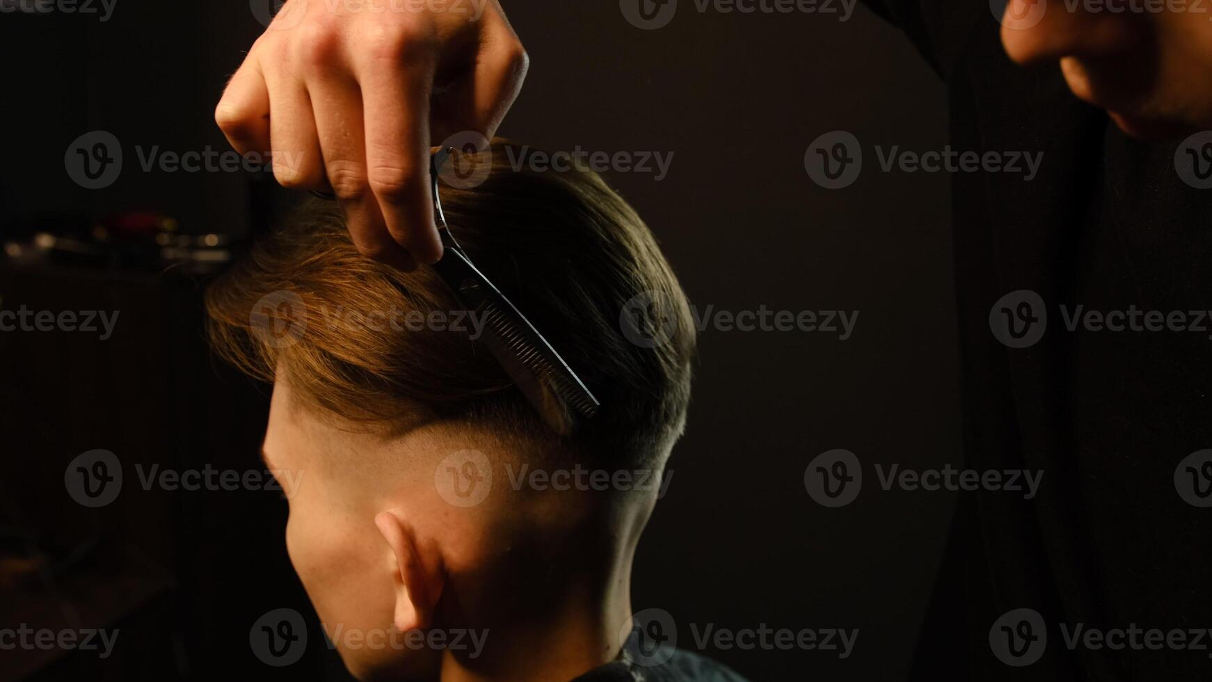 Barber use Thinning scissors and metal comb on brown wavy hair of young man. Hairdresser service in a modern barbershop in a dark key lightning with warm light photo