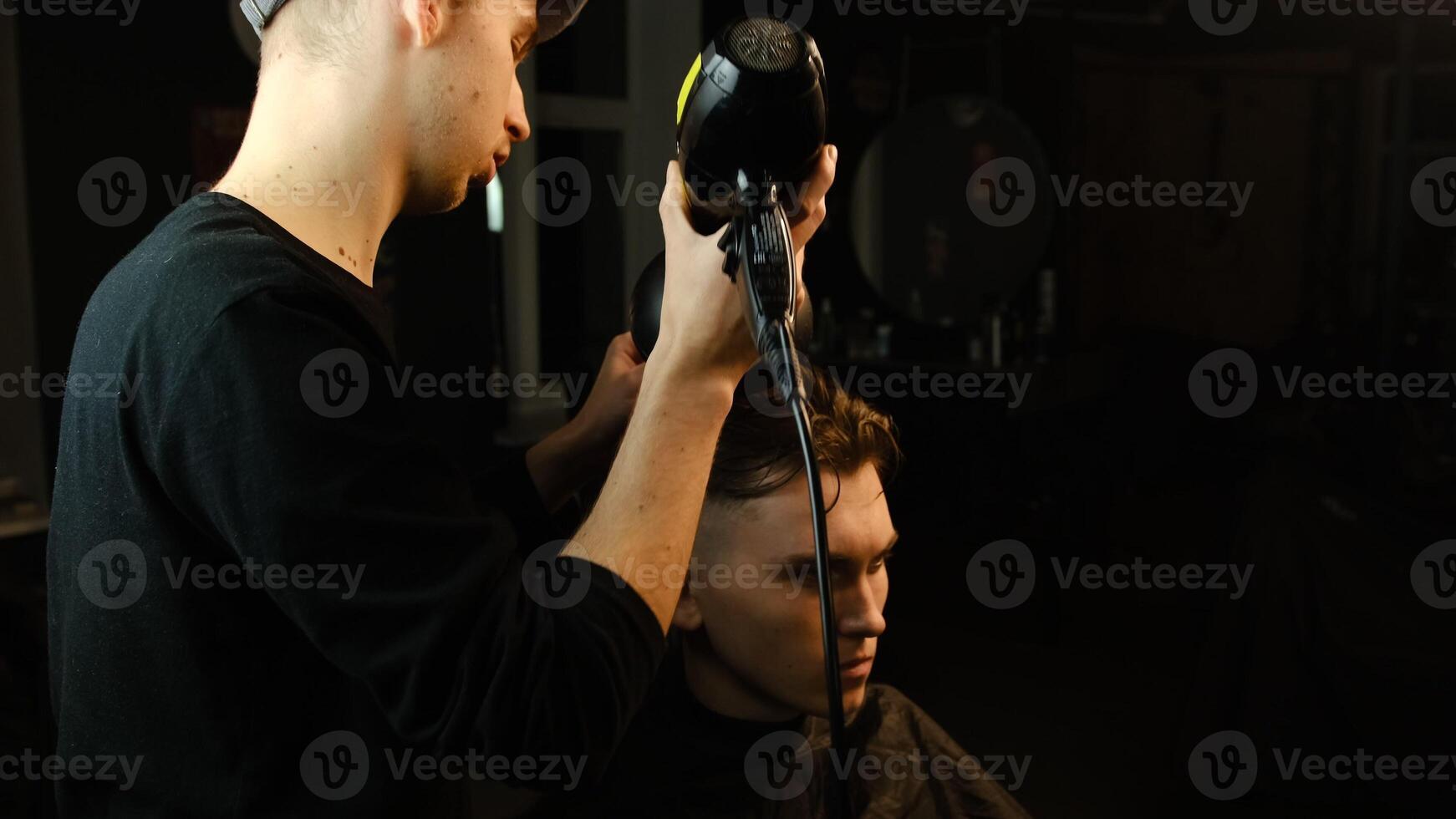 Young barber drying hair of customer with hair dryer in barber shop in a dark key lightning with warm light and correct hair with hand. Hairdresser for man photo