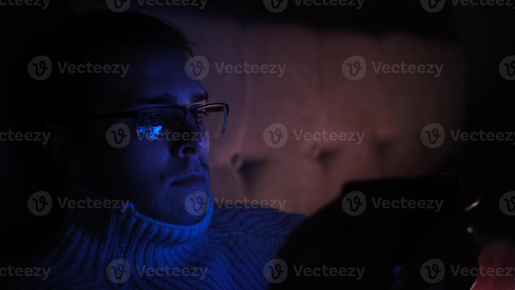 Lonely man in glasses sitting on bed in bedroom and using phone at night. Face illuminated from smartphone display. Surfing social network before sleep photo