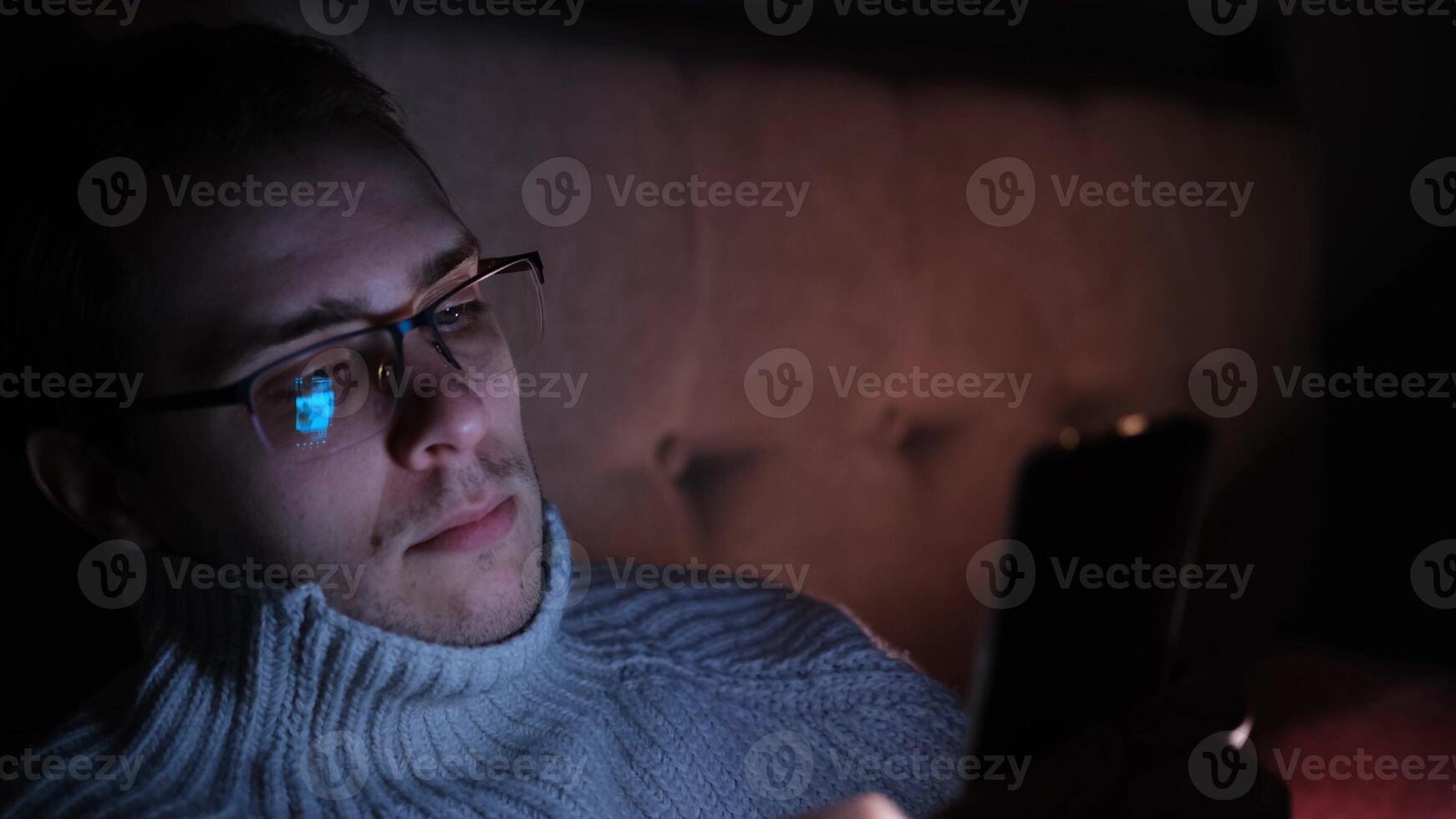 Lonely man in glasses sitting on bed in bedroom and using phone at night. Face illuminated from smartphone display. Surfing social network before sleep photo