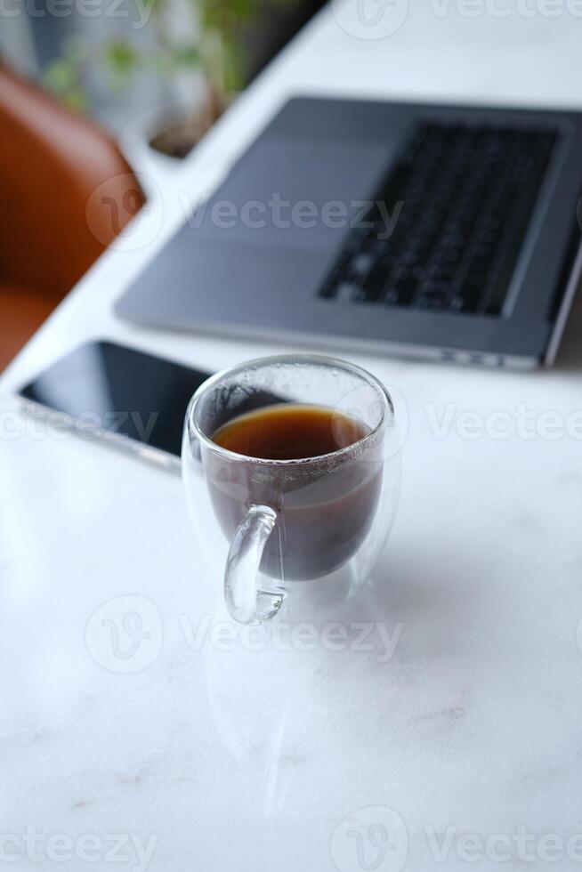 workplace with phone laptop and transparent cup of coffee in home office on white marble desk with brown leathern chair and green tree at background photo