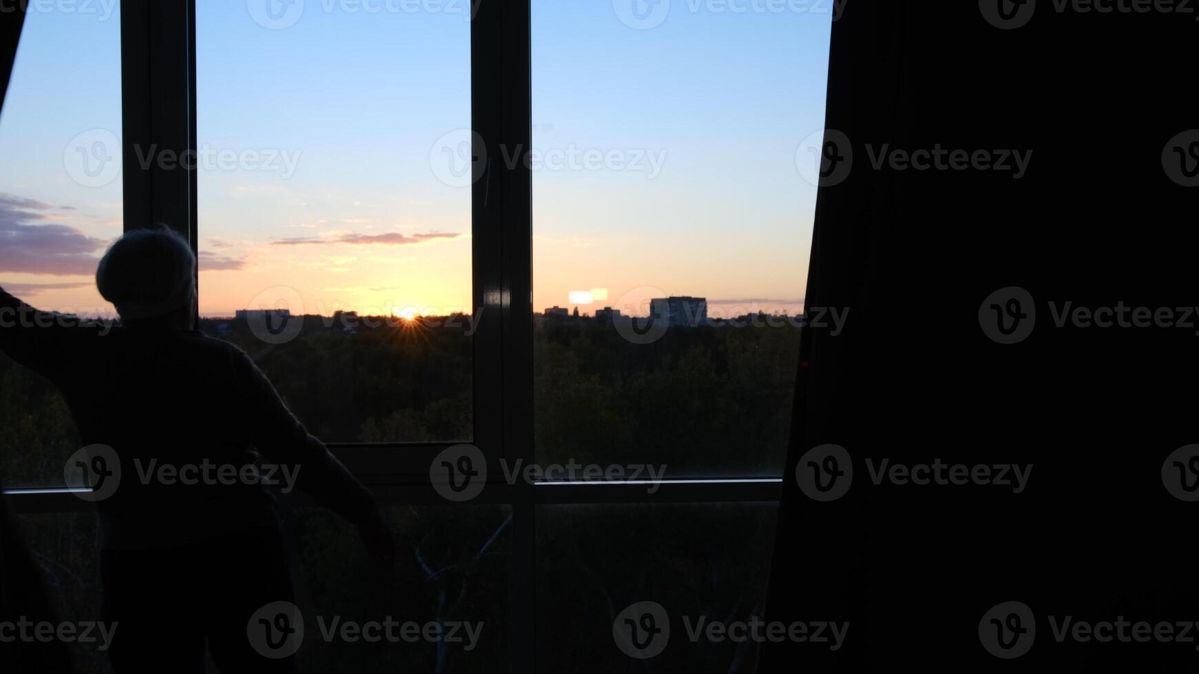 Back view of senior, retired grey haired European woman in sweater letting sunset rays in opening window curtains to look at weather outside before going to work photo