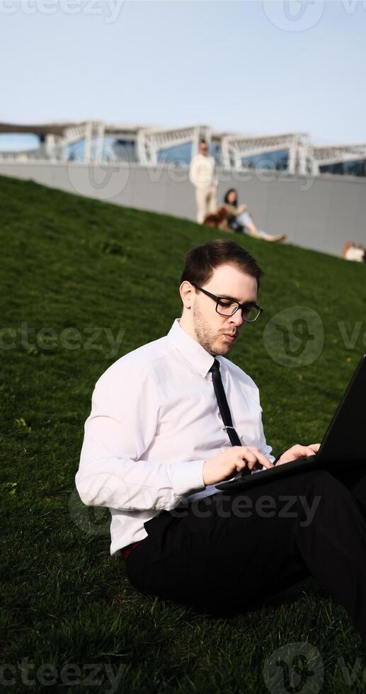 Caucasian bearded business man freelancer working on break with laptop outdoors in park. Male office worker sitting on a green grass on a city park on a modern urban background Workplace outside photo