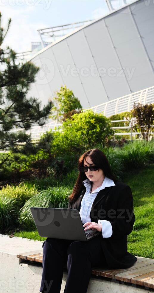 Young business woman in suit freelancer working on break with laptop outdoors in park. Female office worker sitting on a bench on a city street park on a modern urban background Workplace outside photo