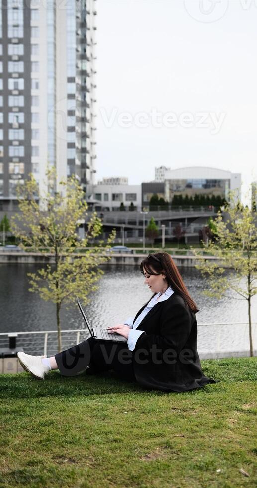 Young business woman in suit freelancer working on break with laptop outdoors in park. Female office worker sitting on a grass on a city street park on a modern urban background Workplace outside photo