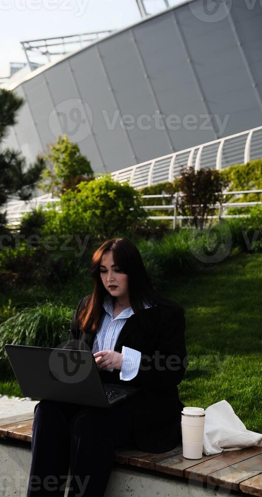 A cheerful young Caucasian woman, either a business professional or student, engages in a hybrid work meeting or online class, presenting with a laptop in a city park. The scene is vertical. photo
