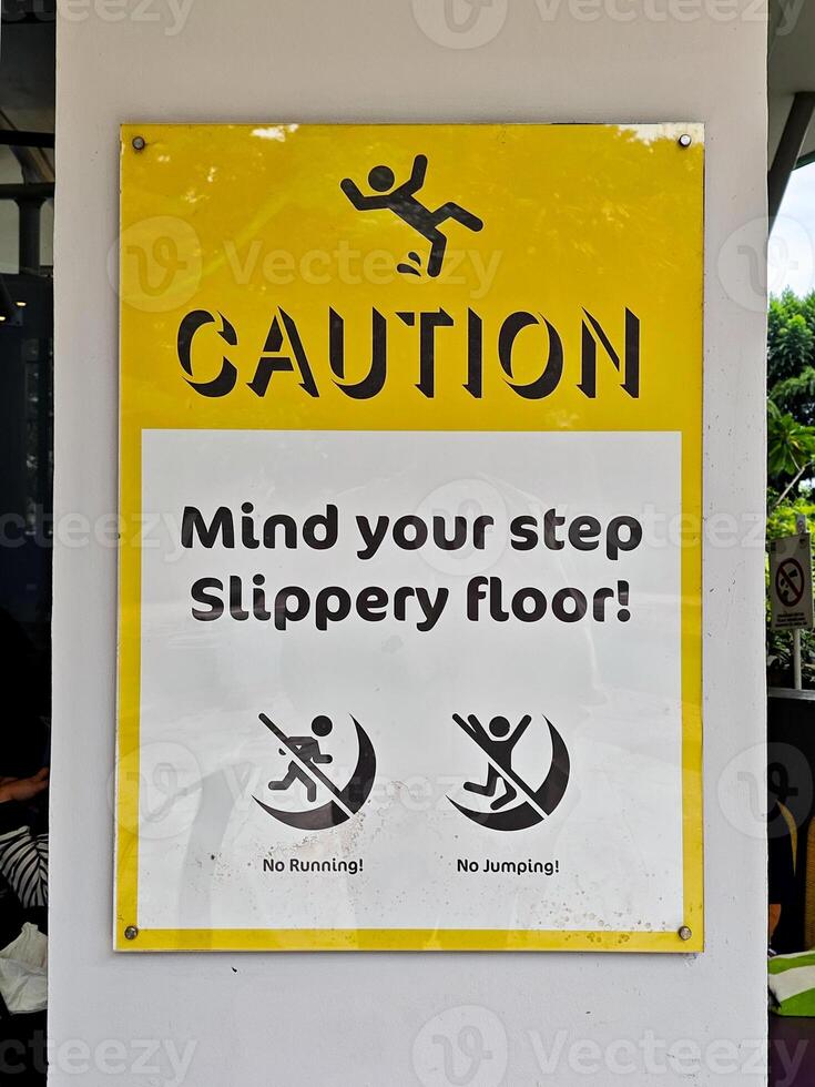 Bright yellow caution sign with stick figure and text Mind your step Slippery floor posted on a swimming pool area emphasizing safety precautions and warning against running or jumping. photo