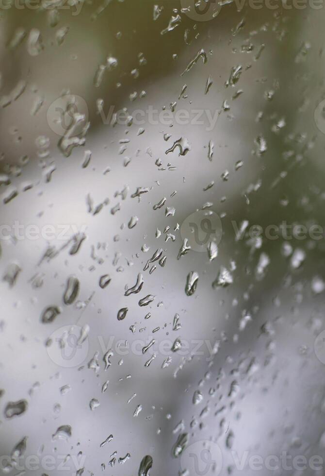 A photo of rain drops on the window glass with a blurred view of the blossoming green trees. Abstract image showing cloudy and rainy weather conditions