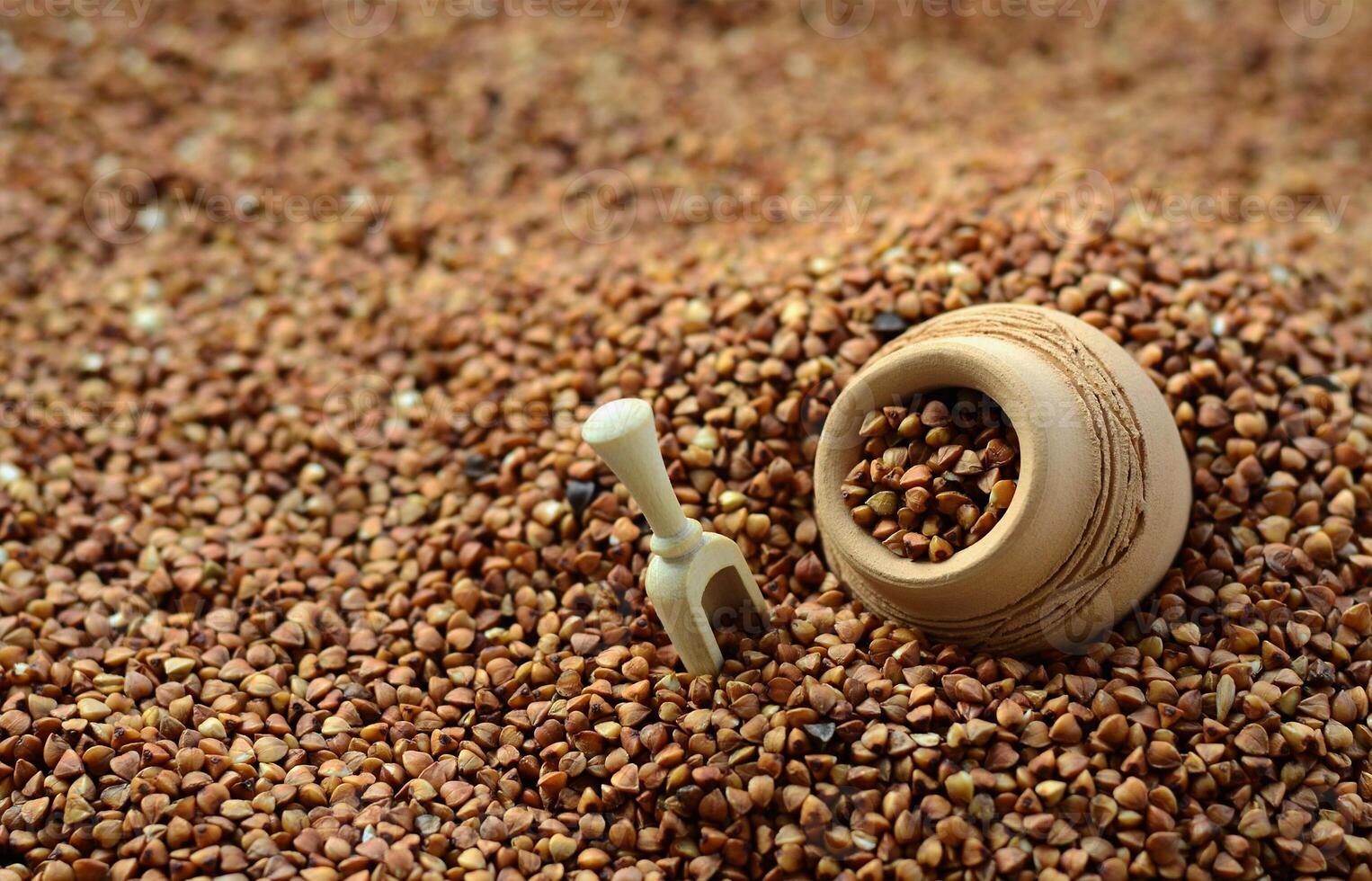 Background image of a large pile of buckwheat, in the middle of which lies a small jug and a wooden spatula for cereals photo