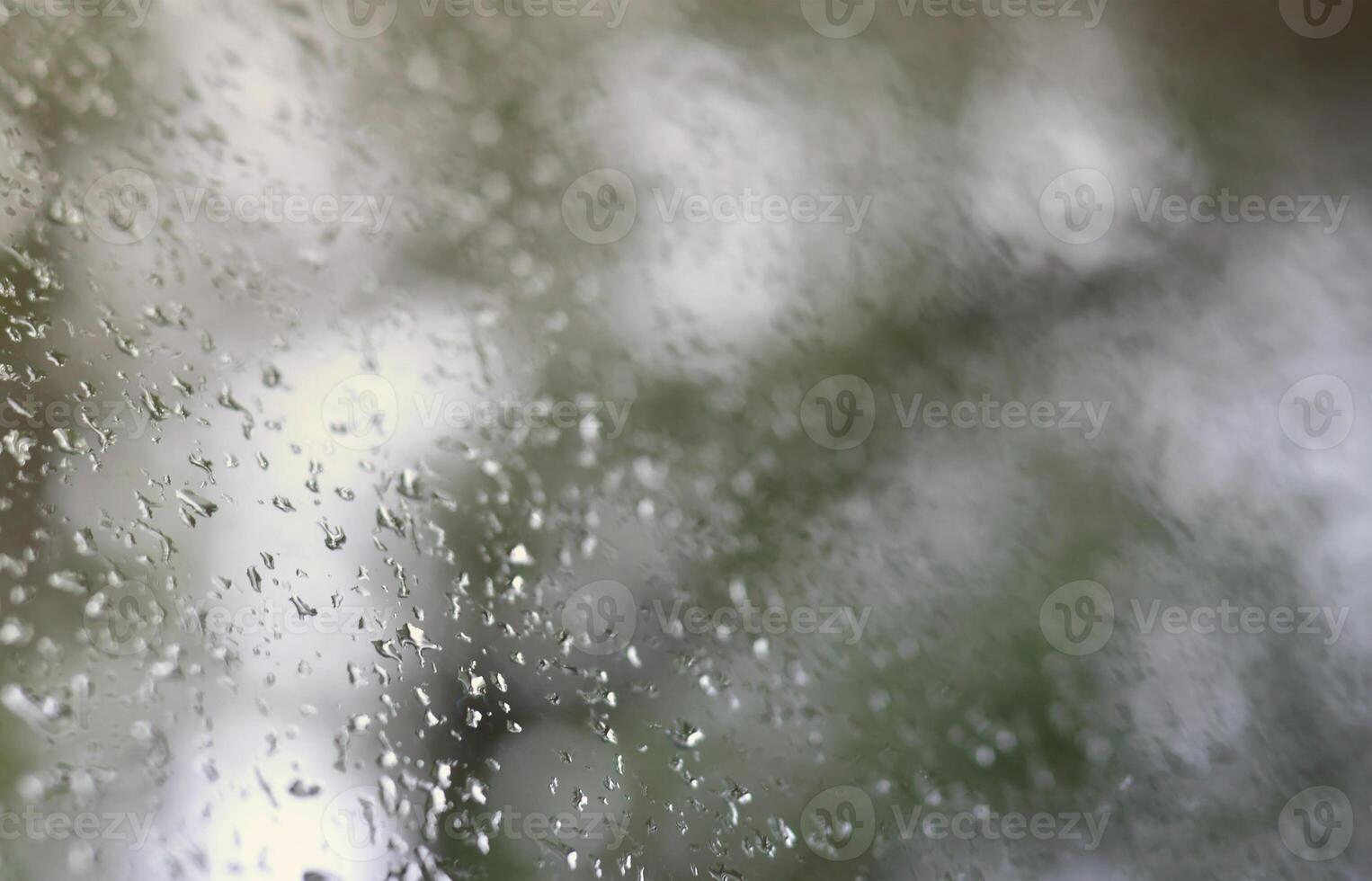 A photo of rain drops on the window glass with a blurred view of the blossoming green trees. Abstract image showing cloudy and rainy weather conditions