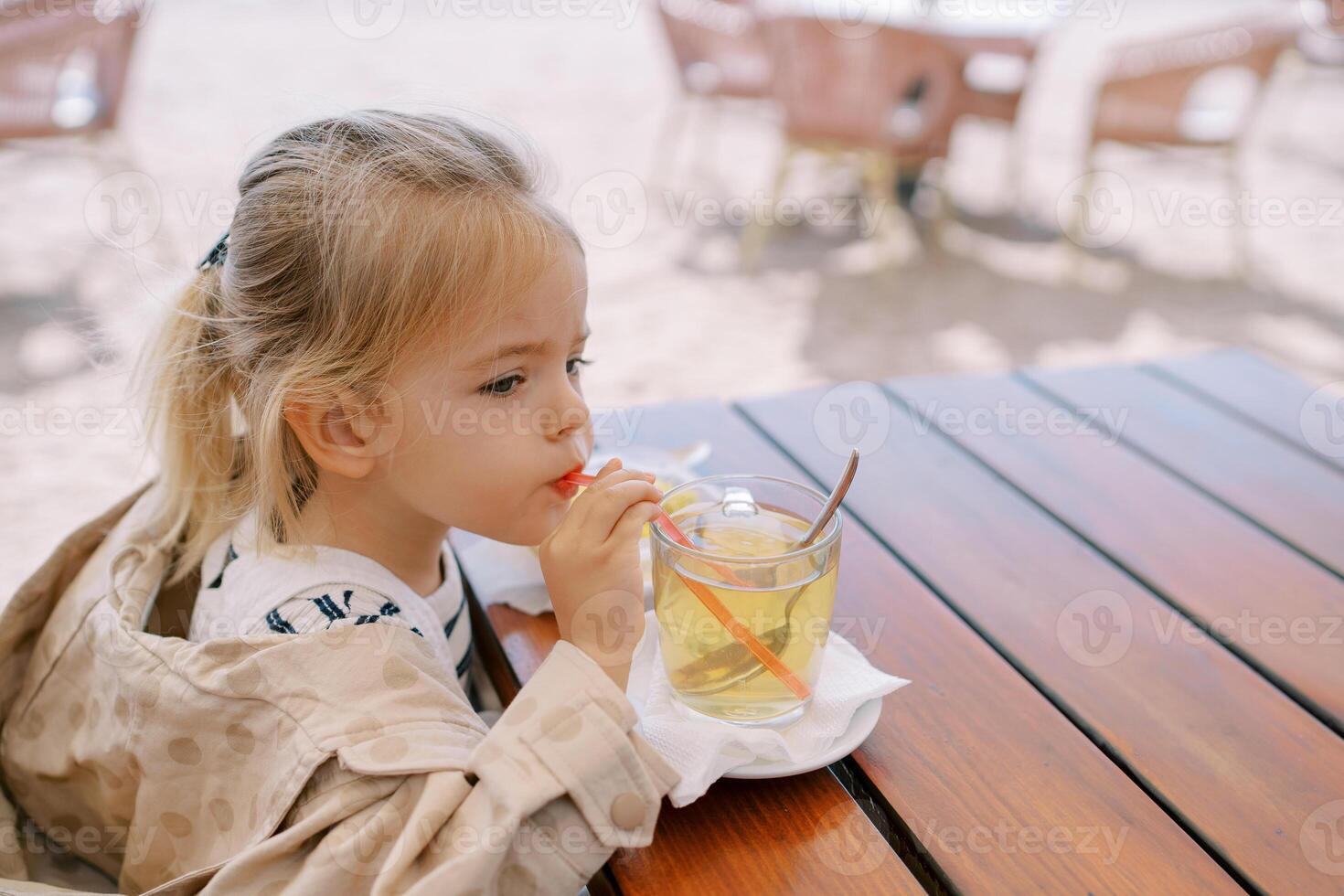 Little girl drinks juice from a glass through a straw while sitting at the table. Side view photo