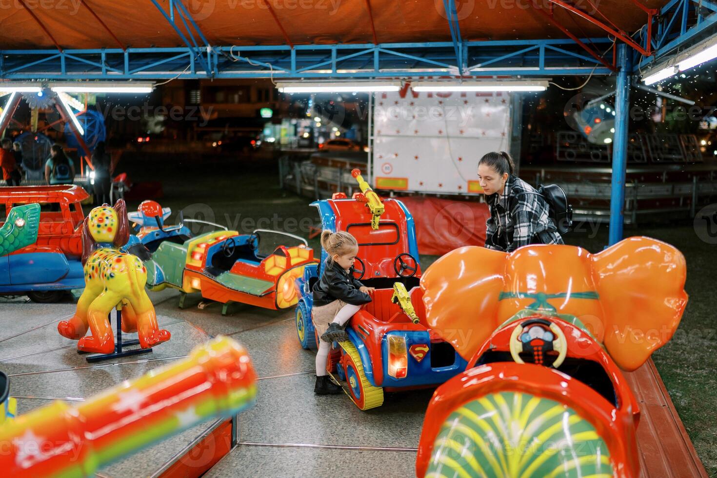 Mom stands near a toy car on a carousel into which a little girl climbs photo