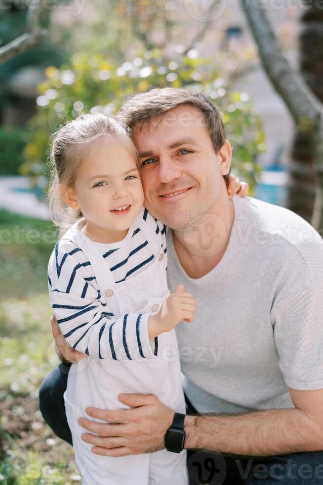 Little girl hugs the neck of her smiling dad squatting in the garden photo