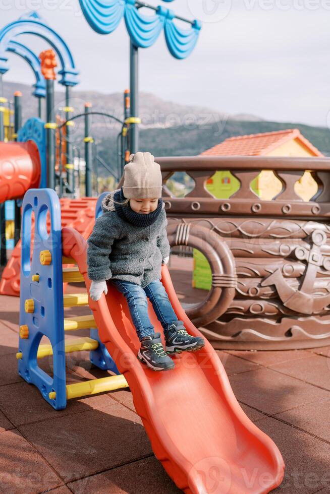 Little smiling girl in mittens slides down a slide on the playground photo