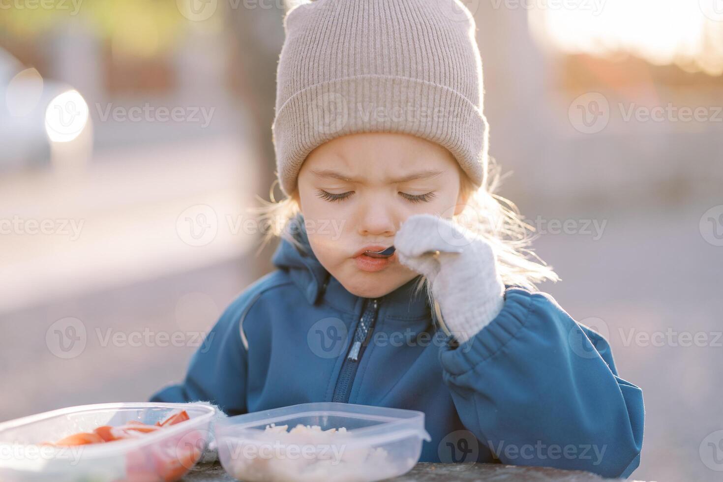 Little girl eats porridge with a spoon looking into her lunchbox while standing in the park photo
