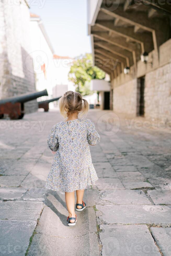 Little girl walks through the courtyard of an ancient castle towards the cannons standing against the wall. Back view photo