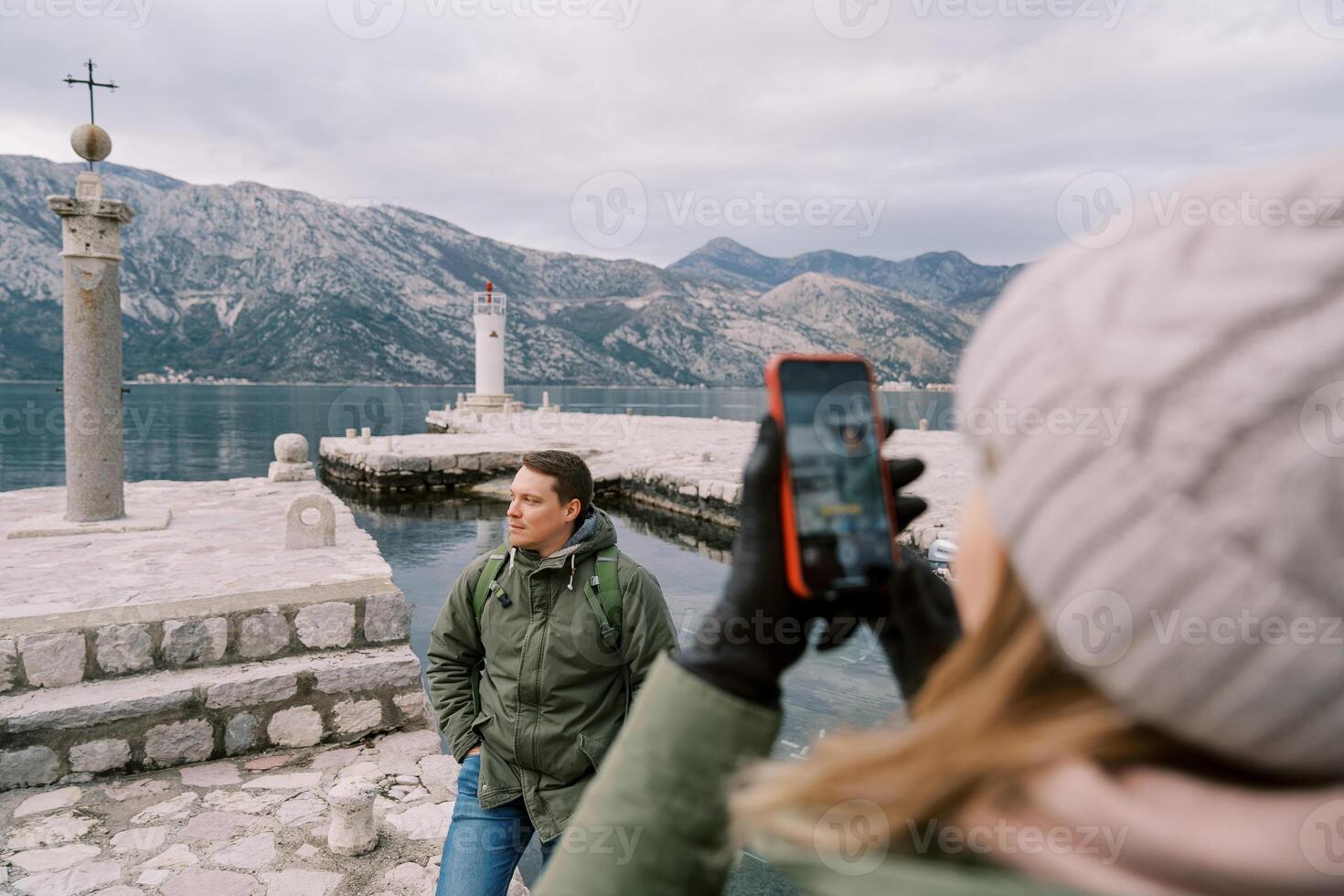 Woman takes a picture with a smartphone of a man standing on a pier near a column with a cross. Back view photo