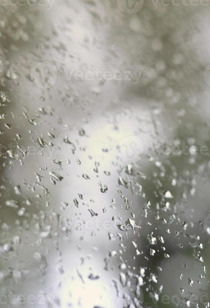 A photo of rain drops on the window glass with a blurred view of the blossoming green trees. Abstract image showing cloudy and rainy weather conditions