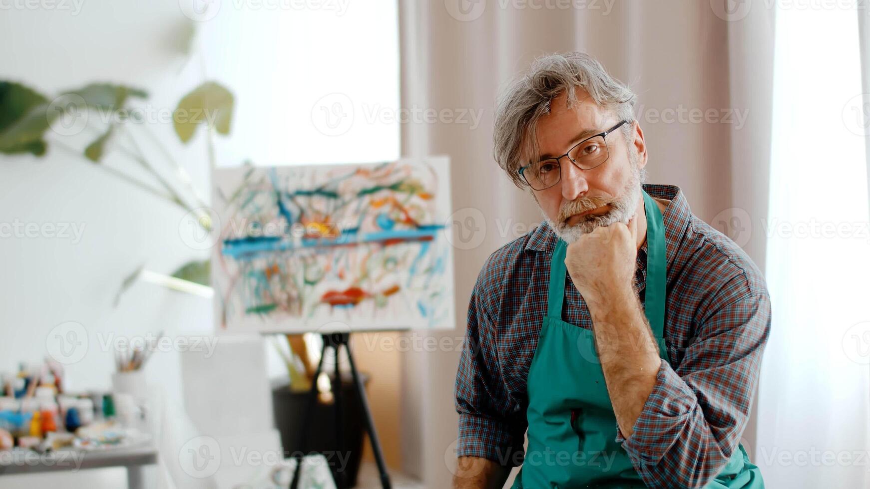 Man Artist In An Apron Sits Thoughtfully On Chair In Studio And Looks At Camera photo