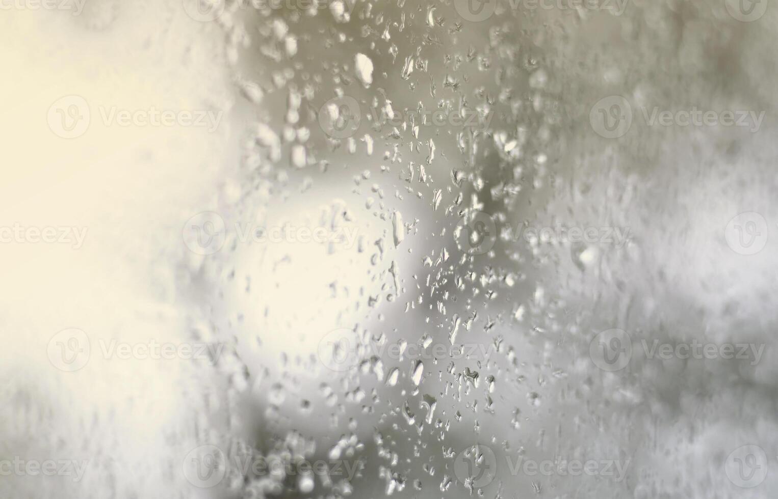 A photo of rain drops on the window glass with a blurred view of the blossoming green trees. Abstract image showing cloudy and rainy weather conditions