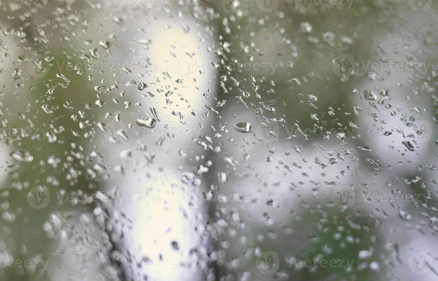 A photo of rain drops on the window glass with a blurred view of the blossoming green trees. Abstract image showing cloudy and rainy weather conditions