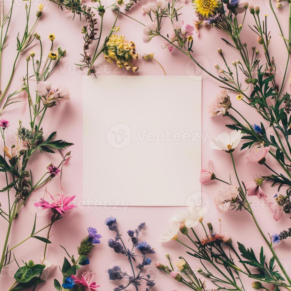 Square blank card surrounded by pastel colored flowers on a light pink background in a top view photo