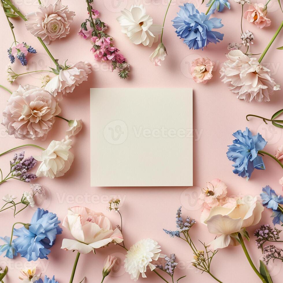 Square blank card surrounded by pastel colored flowers on a light pink background in a top view photo