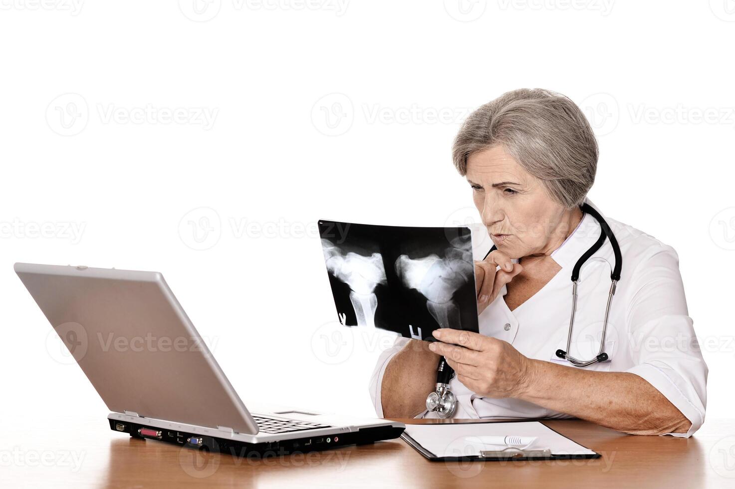 Serious old woman doctor sitting at table with computer and looking at x-ray photo