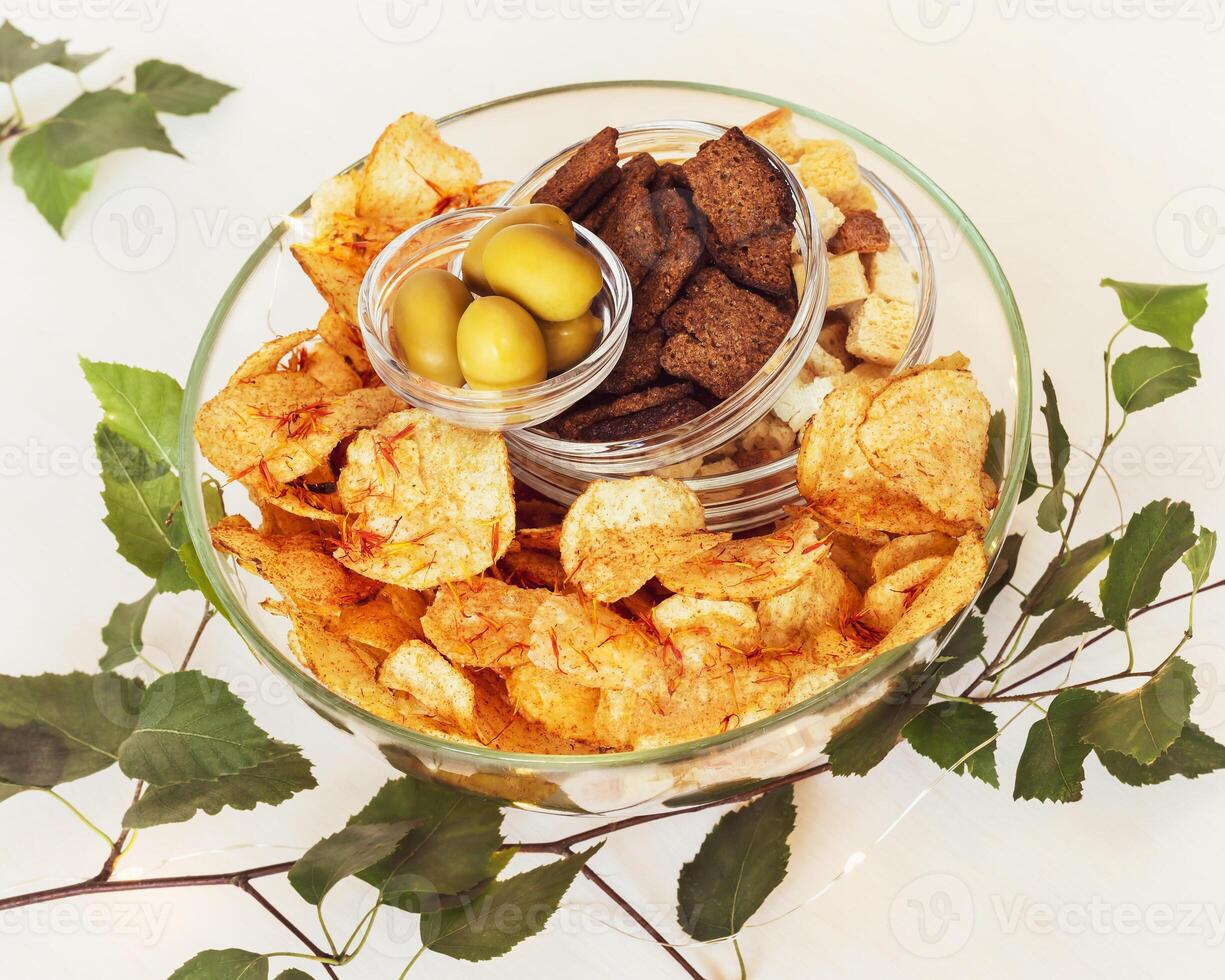 Rye crackers, wheat crackers, potato chips with spices and green olives in glass bowls on a white table photo