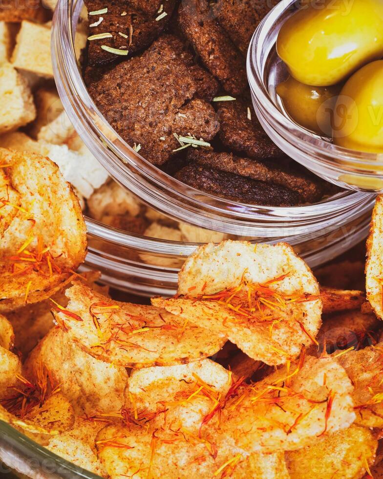Rye crackers, wheat crackers, potato chips with spices and green olives in glass bowls on a white table photo