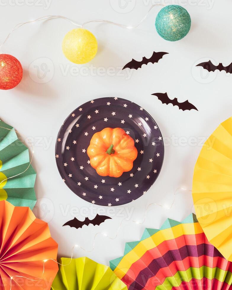 Small pumpkin on a plate, colorful paper fans, helloween decorations and light garland on a blue background photo