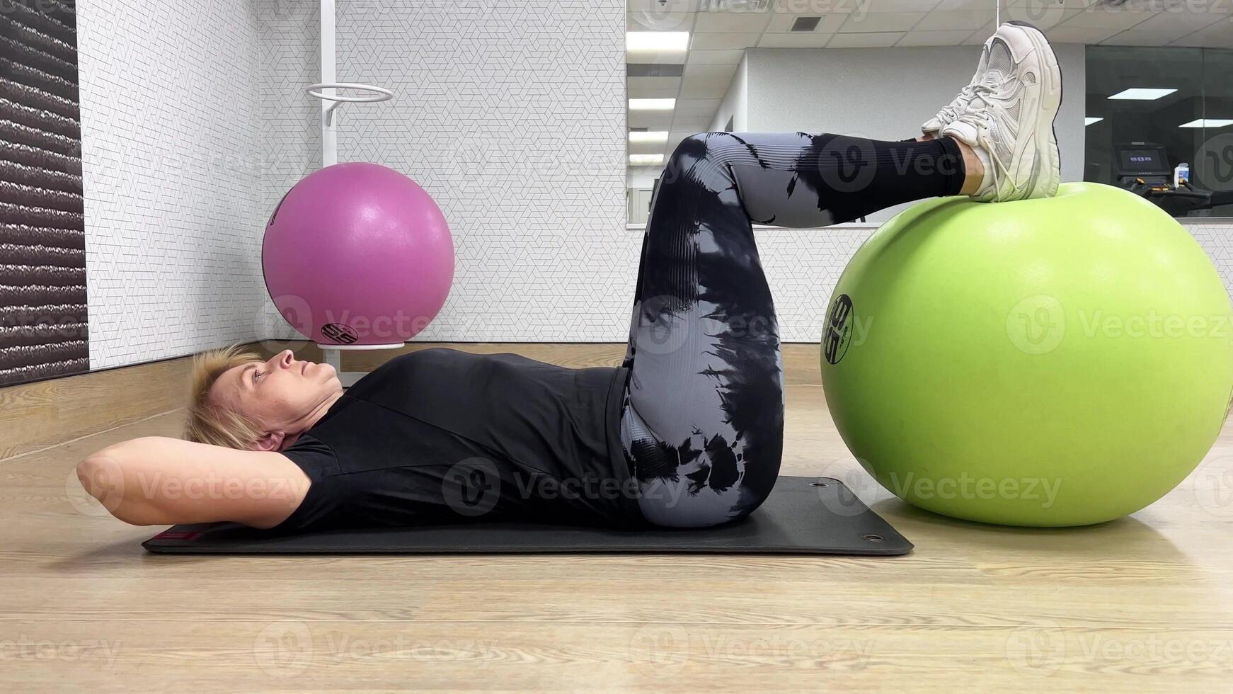 Woman 55-60 years in the gym. A woman lies on her back with her hands behind her head and her feet on a fitness ball. A woman performs exercises with a fitness ball. photo