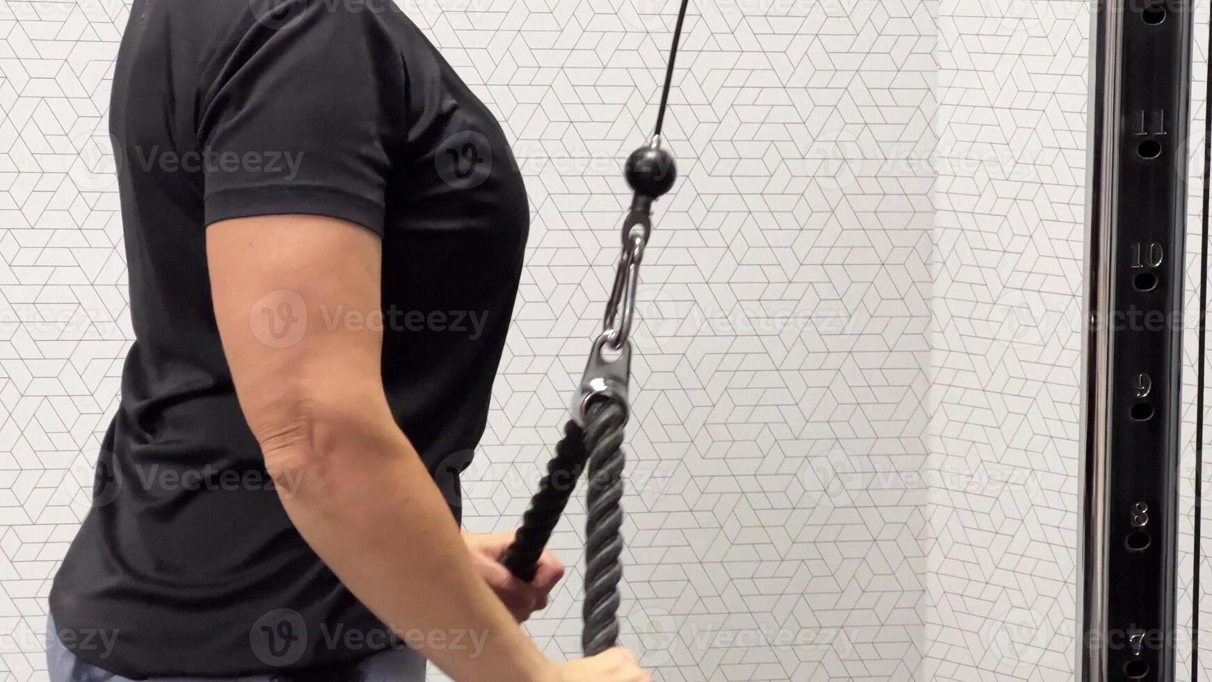 A person in a gym, wearing a black t-shirt, is pulling on a cable machine for resistance training. photo