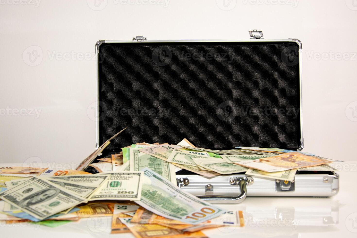 A silver briefcase is open with money spilling out onto a white surface photo