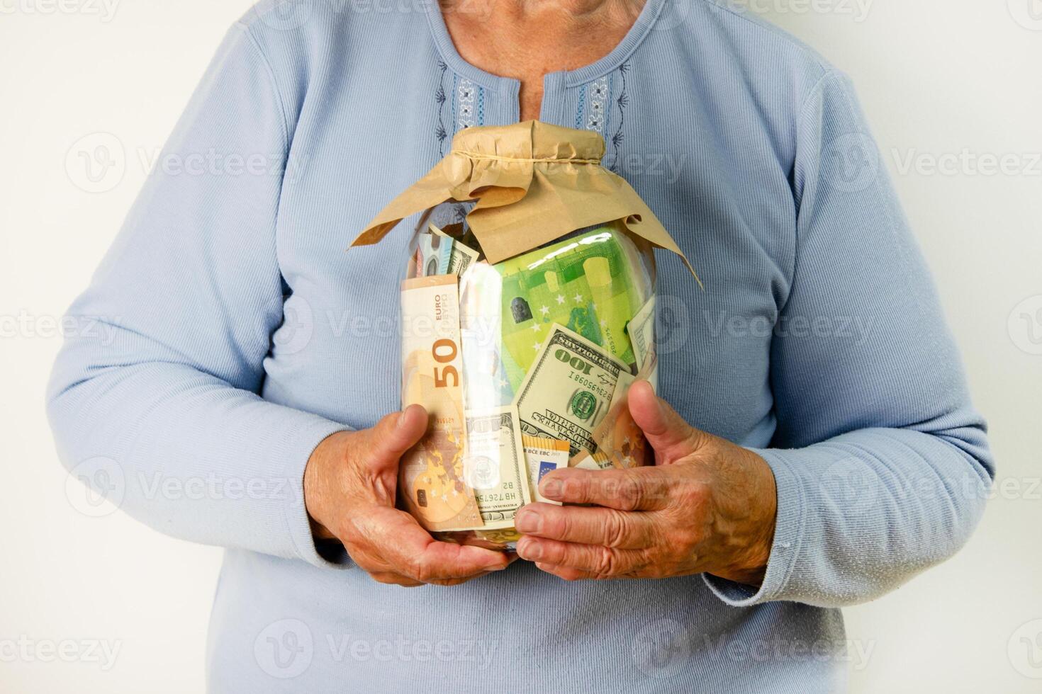 An elderly woman holds a glass jar filled with a mixture of dollar bills and euro banknotes in front of a white background photo