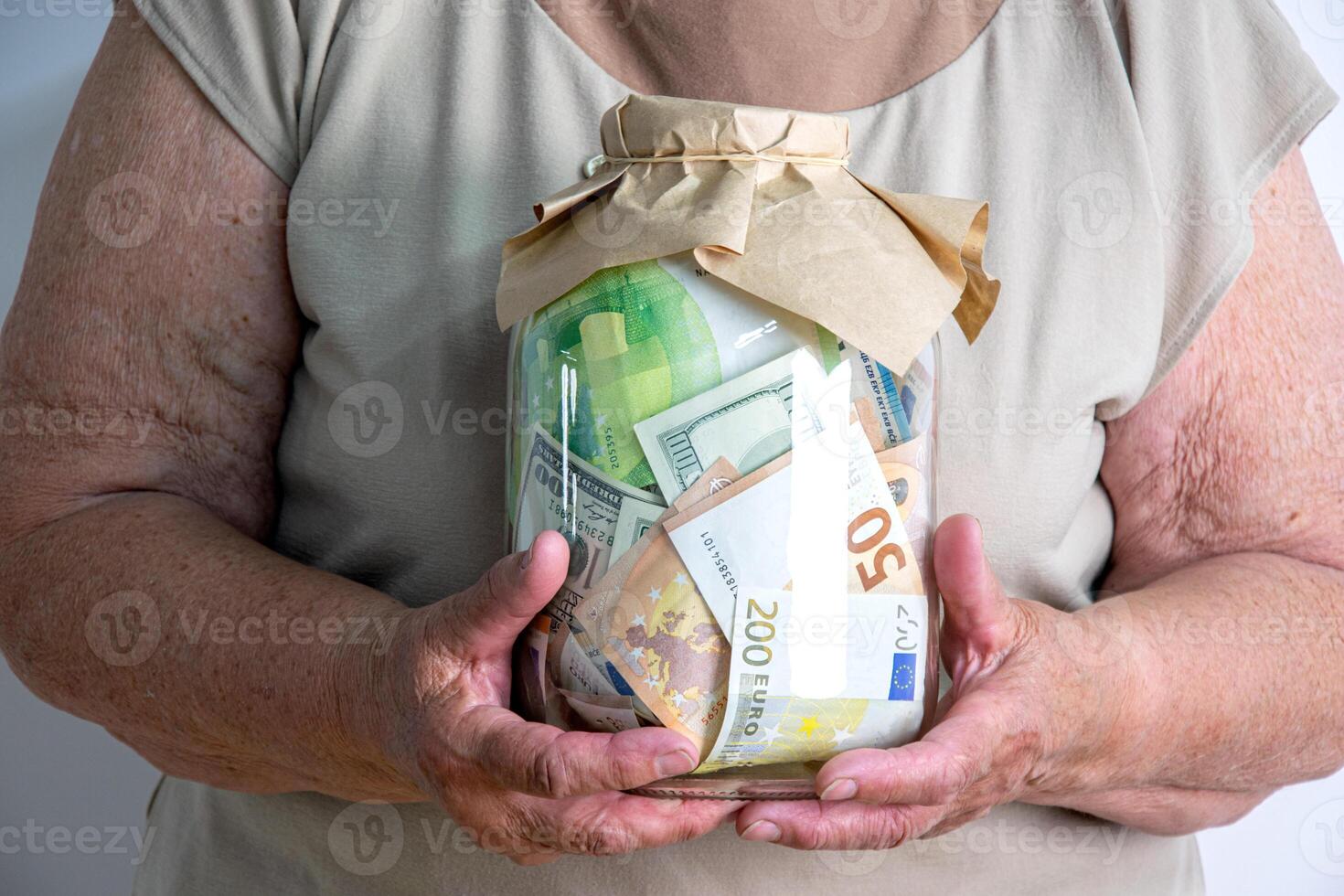 An older woman holds a glass jar filled with euro and dollar bills in her hands photo