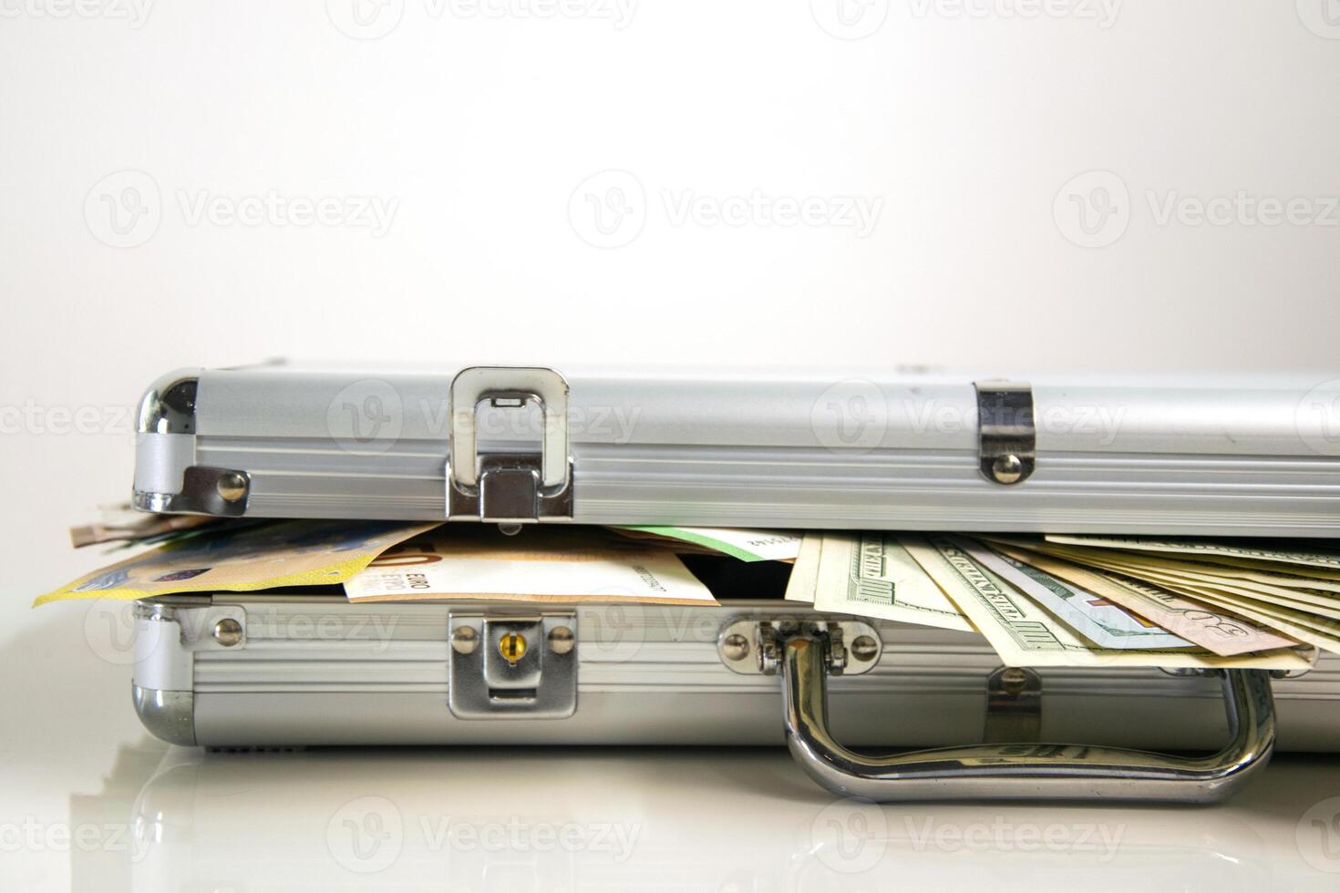 A close-up of an open silver aluminum case with cash spilling out from inside. The case is on a white surface with the cash slightly overlapping the edge photo