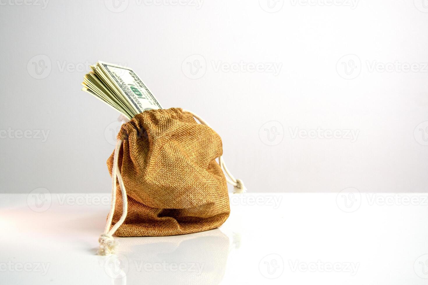 A brown canvas bag with a drawstring sits on a white surface. Several US dollar bills are sticking out of the top of the bag photo