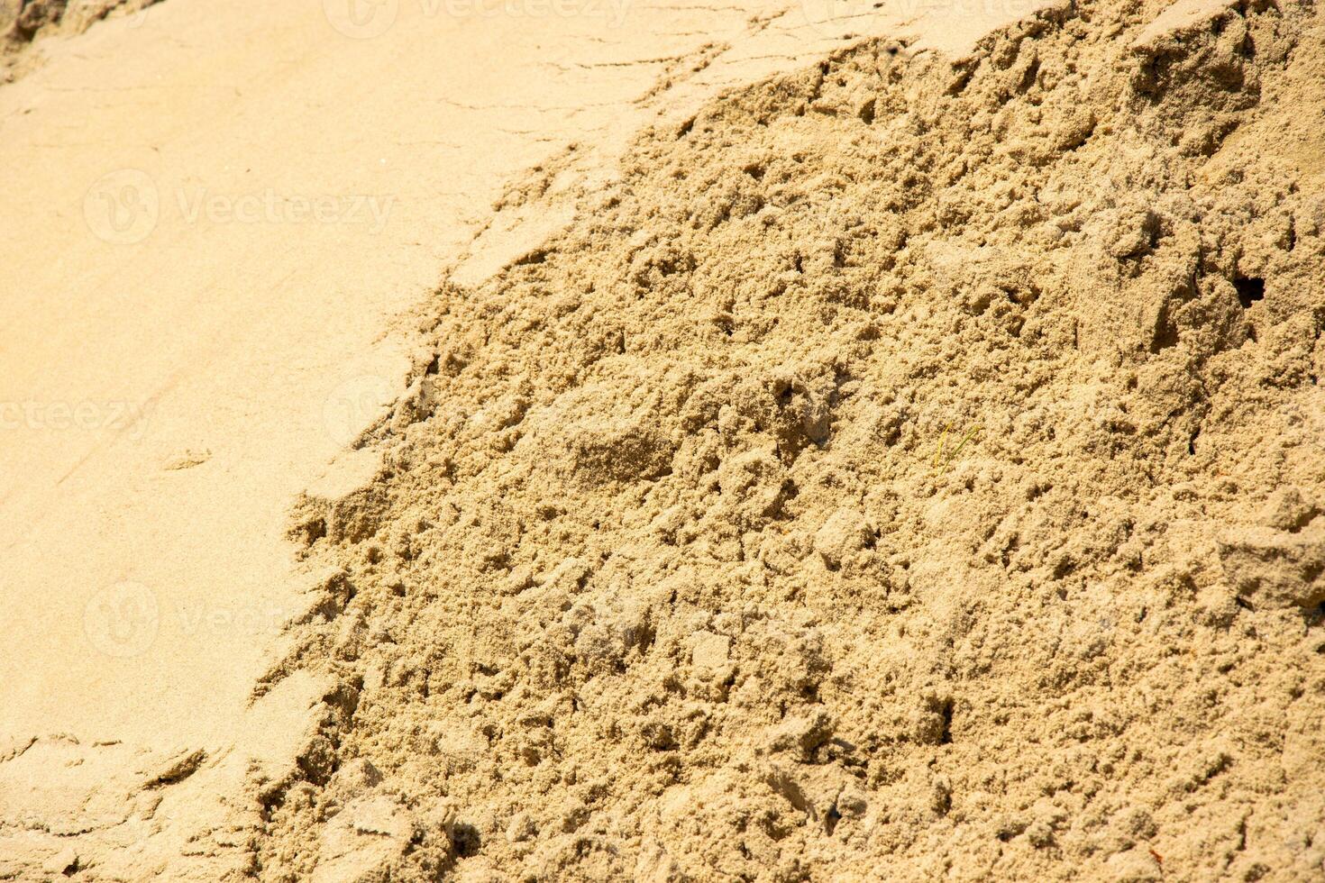 A close-up image of a pile of light brown sand, likely taken outdoors on a sunny day. The sand appears to be dry and loose, with a coarse texture photo