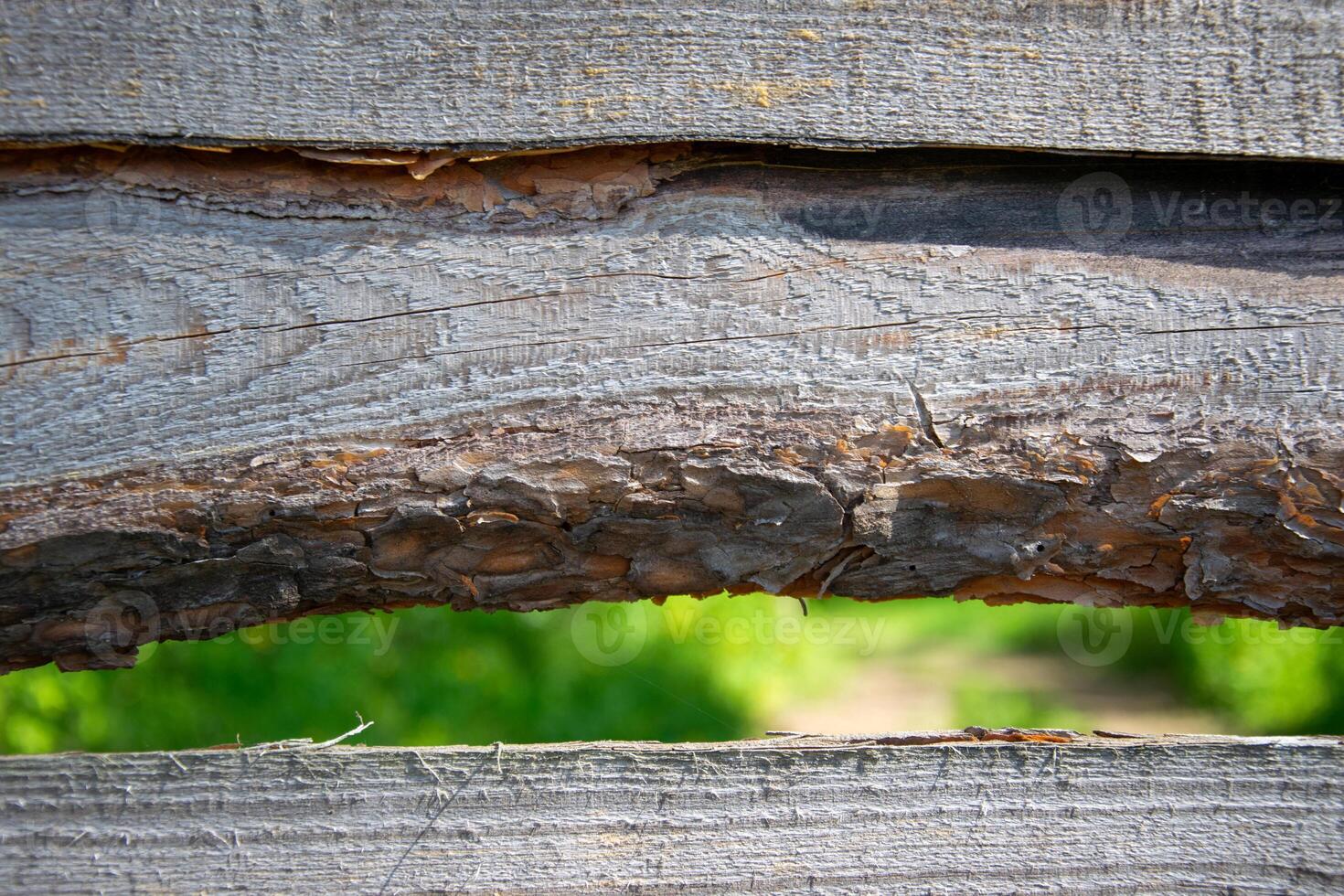 A close-up view of a weathered wooden fence with a small gap revealing a blurred green landscape photo