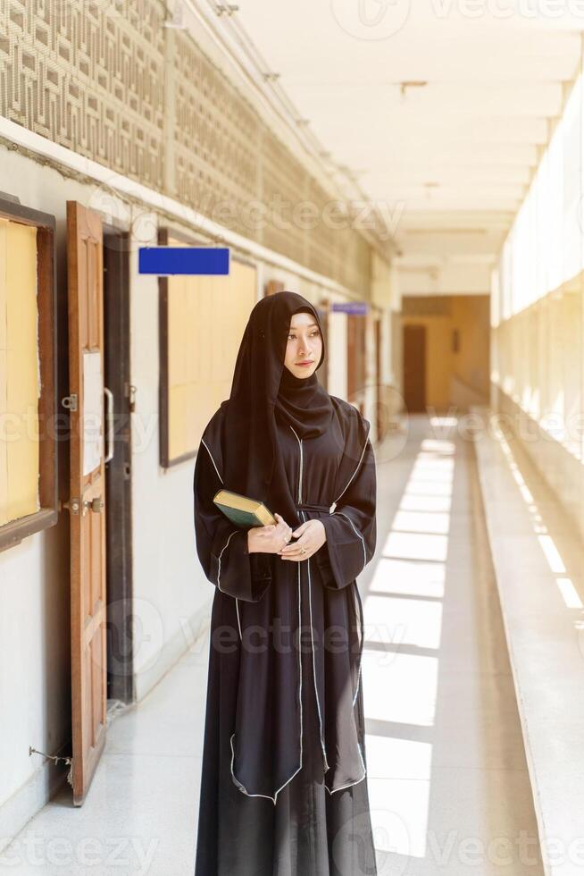 A Muslim student wearing traditional clothes standing and hold Quran on walkway in her school building with sun flare background. photo