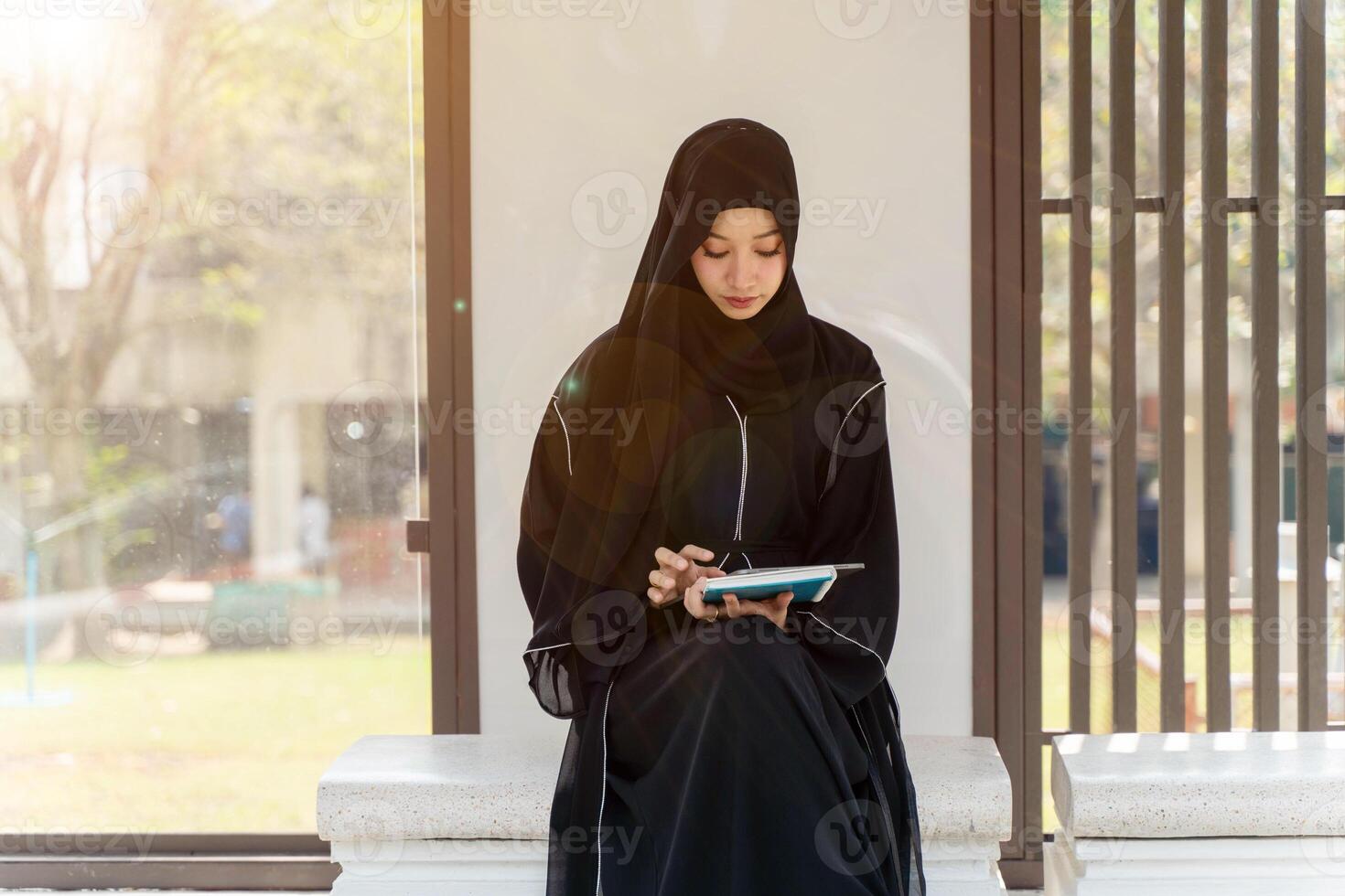 Asian Muslim wearing traditional clothes sit and review the lesson on tablet for the exam in university with sun fare and blurred background. photo