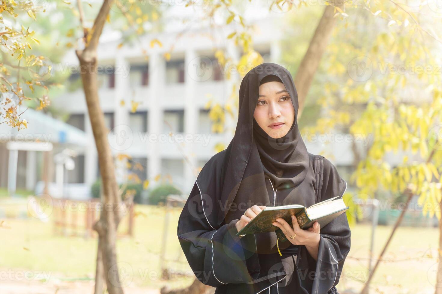 Closeup and crop Muslim student wearing traditional clothes stand and holding Quran on her school building with sun flare background. photo