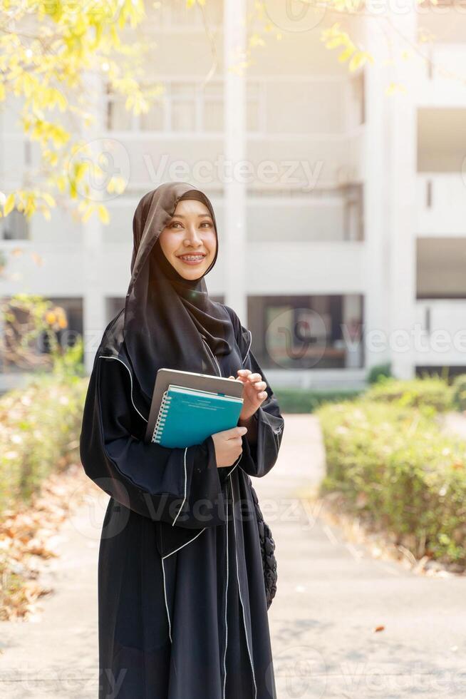 Cheerful Asian Muslim wearing traditional clothes stand and holding textbook on walkway on her school building with sun flare background. photo
