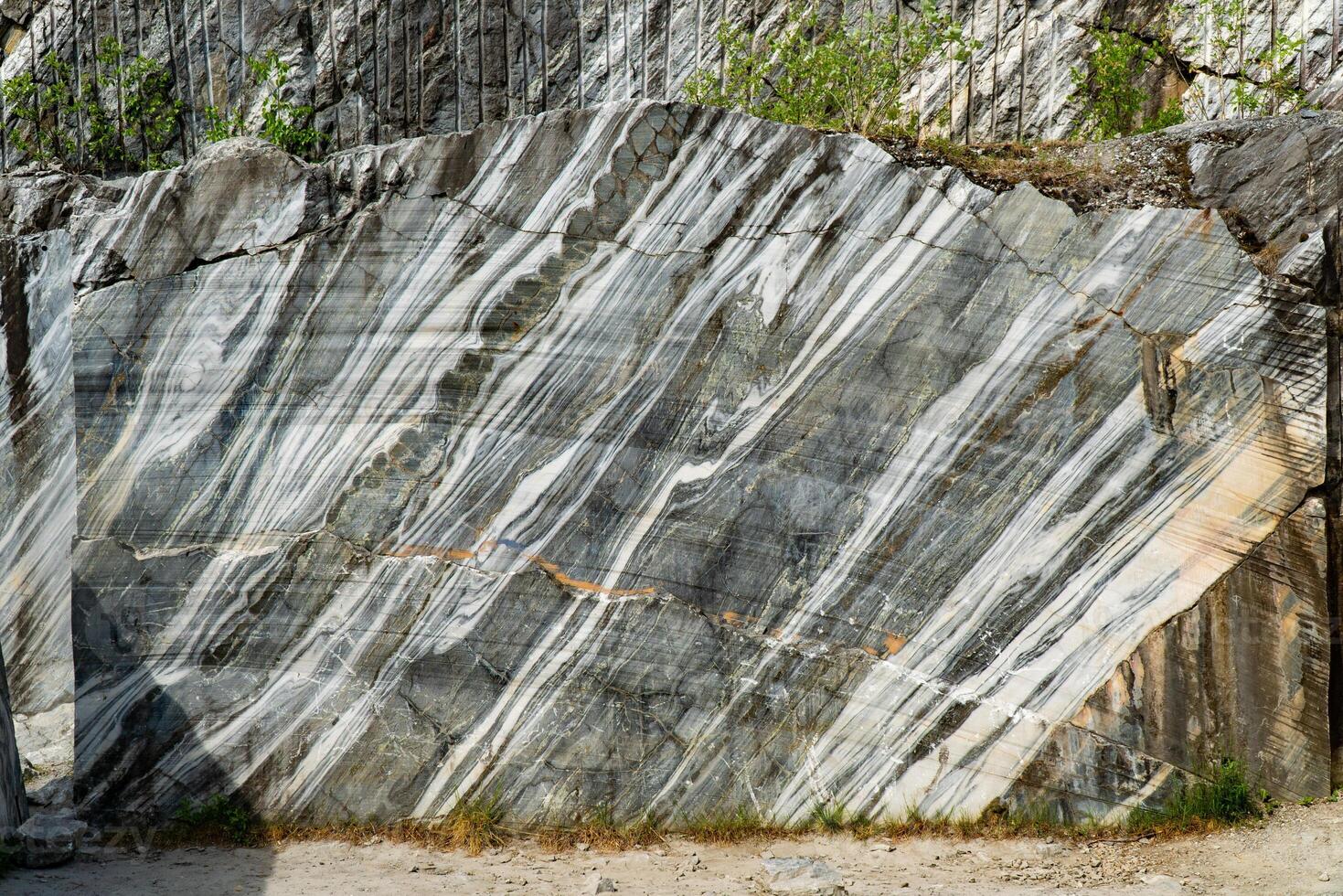 Scenic view of a marble quarry with distinct striations and pine trees in the background. The image highlights the contrast between the natural stone formations and the greenery under a bright blue sky. photo
