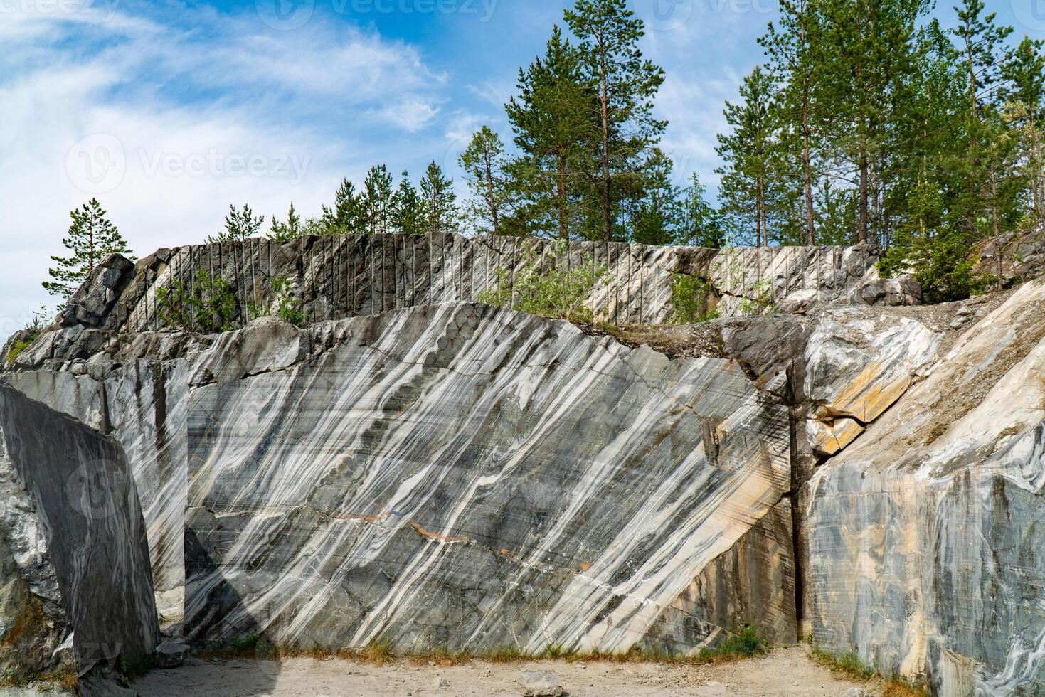 Scenic view of a marble quarry with distinct striations and pine trees in the background. The image highlights the contrast between the natural stone formations and the greenery under a bright blue sky. photo