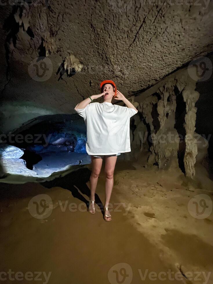 A woman wearing an oversized white t-shirt and a red helmet explores a dimly lit cave, looking up at the stalactites and cave formations with a curious expression. photo